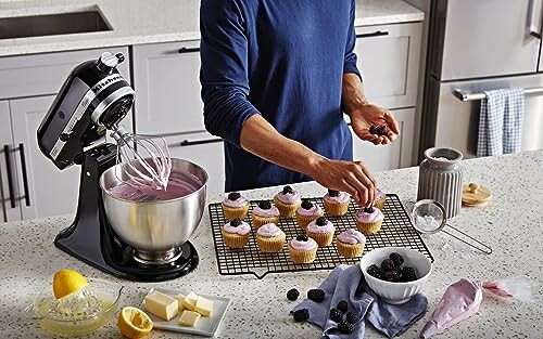 Person decorating cupcakes next to a mixer on a kitchen counter.